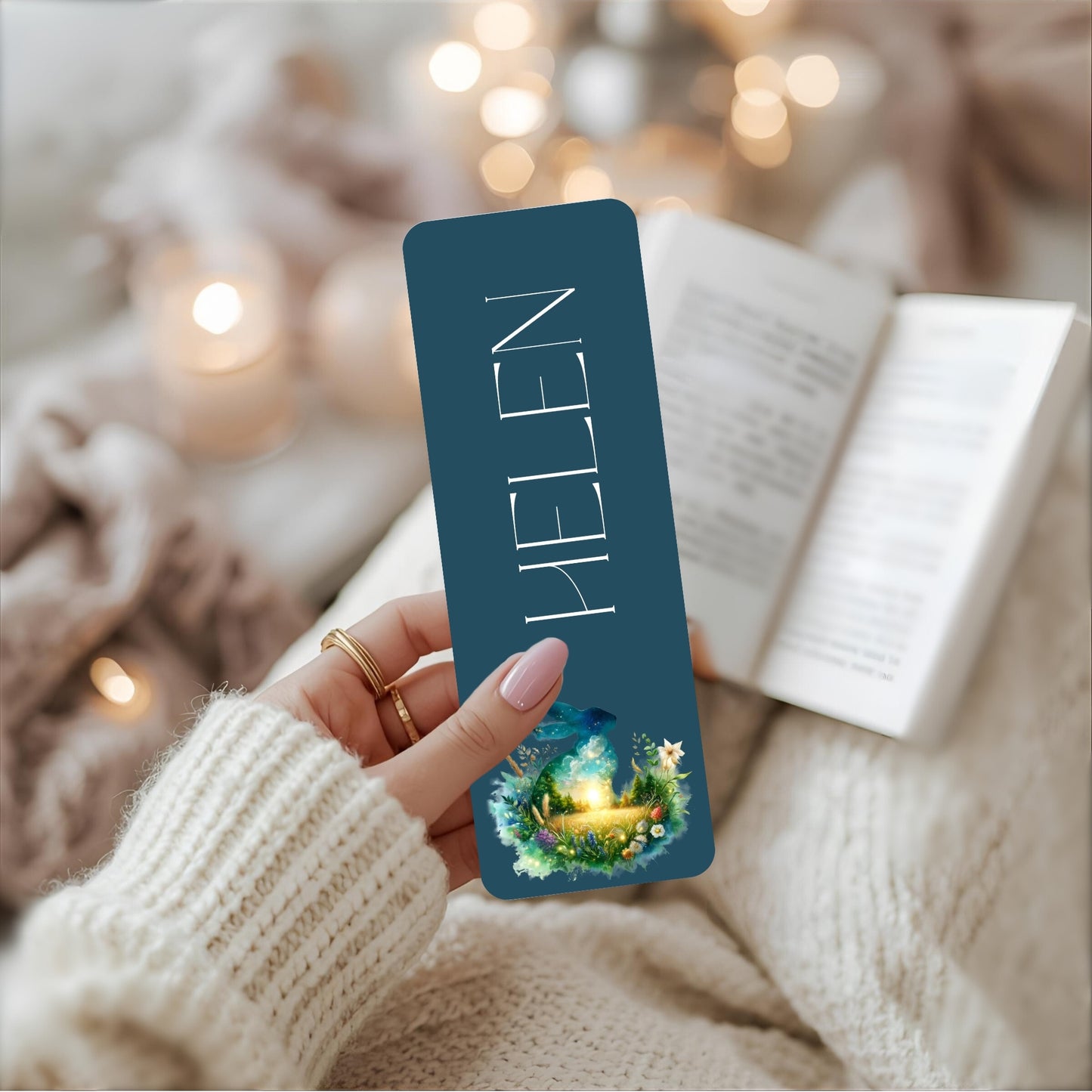 Hand holding a blue bookmark with 'HELEN' and a hare design, against a cosy background.