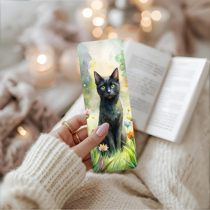 Person holding a bookmark with a black cat design, surrounded by soft lighting and cosy fabric.