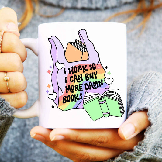 White mug held in hands with a carrier bag with books in illustration, text reads "I work so I can buy more damn books"