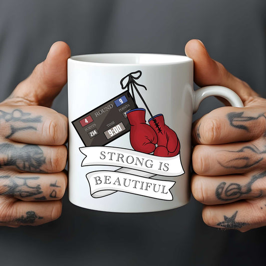 White mug with boxing glove design and 'Strong is Beautiful' text held by tattooed hands against a dark background