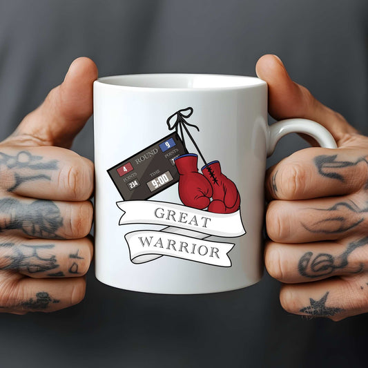 White mug with boxing glove design and 'Great Warrior' text held by tattooed hands against a dark background