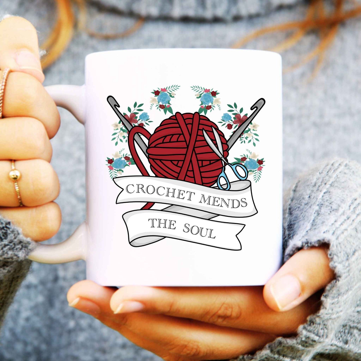 White mug with crochet-themed design held by a person wearing a grey sweater.