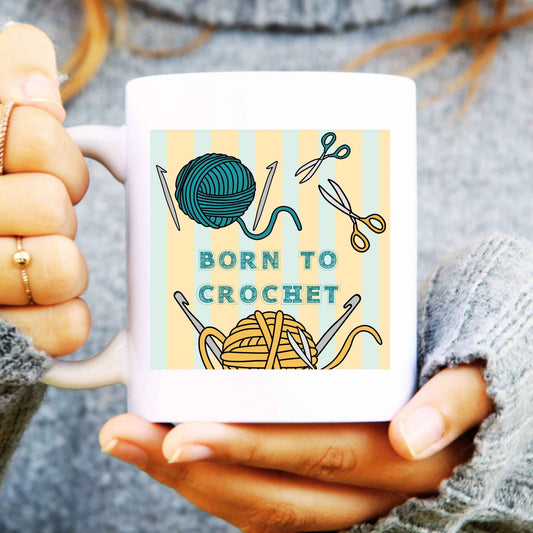 Person holding a mug with 'Born to Crochet' design featuring yarn and crochet hooks.