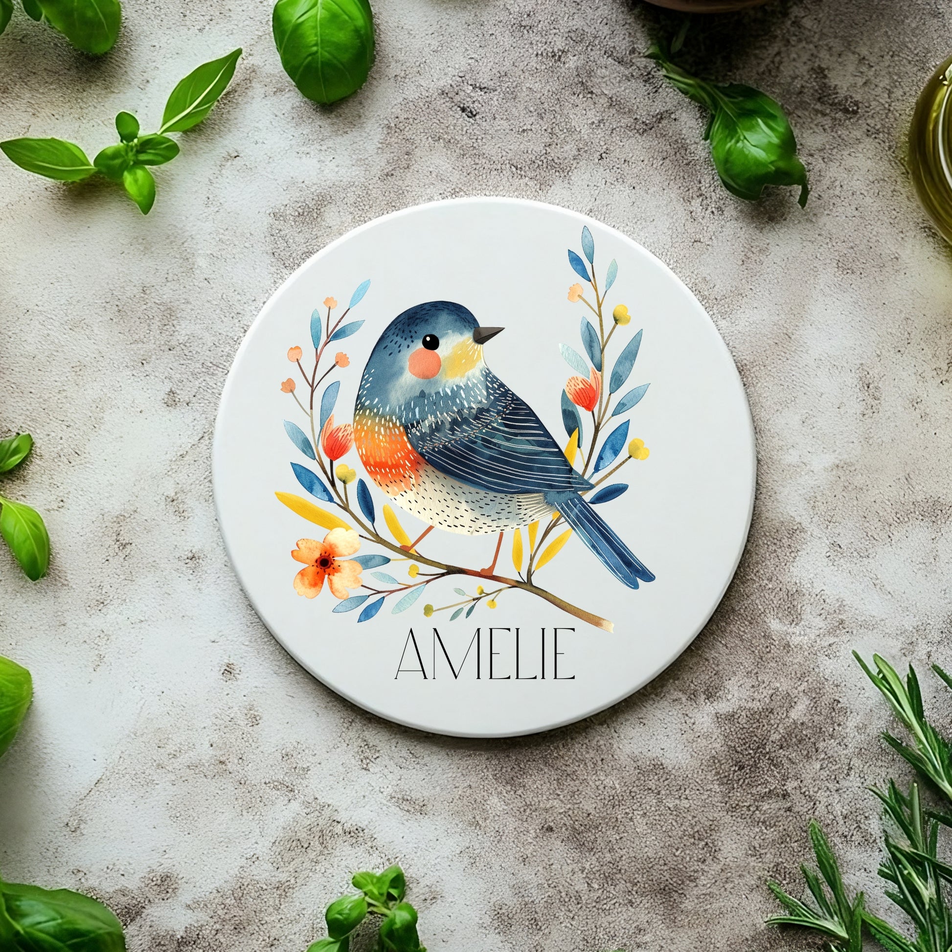 Round ceramic coaster with a bird design and 'Amelie' text, surrounded by green leaves on a textured surface.
