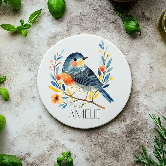 Round ceramic coaster with a bird design and 'Amelie' text, surrounded by green leaves on a textured surface.