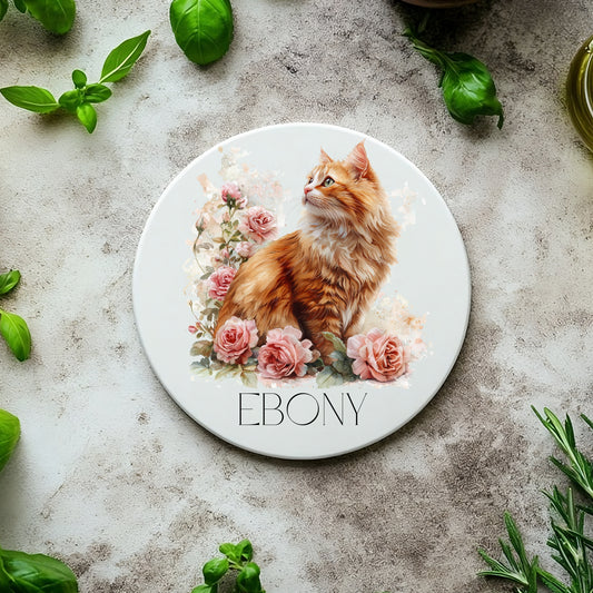 Round white coaster with a cat illustration and the name 'Ebony' on a textured surface with green leaves.