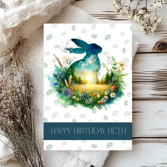 Greeting card with a blue hare illustration and 'Happy Birthday Beth' text, surrounded by flowers on a wooden surface.
