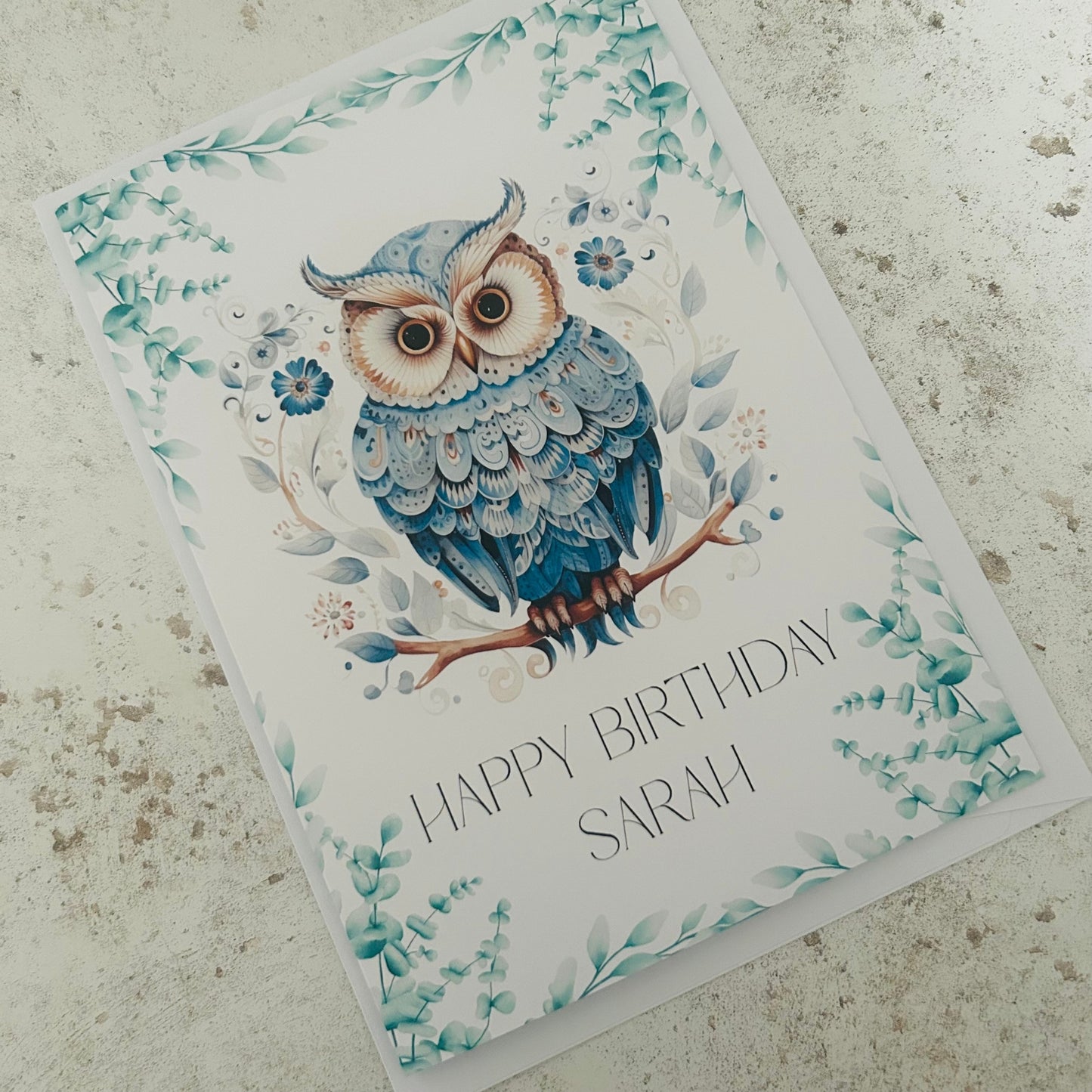 Greeting card with a blue owl illustration and 'Happy Birthday Sarah' text on a textured surface.