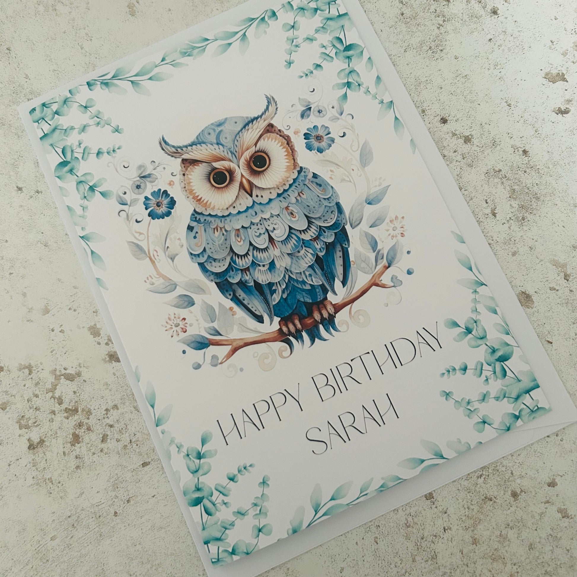 Greeting card with a blue owl illustration and 'Happy Birthday Sarah' text on a textured surface.