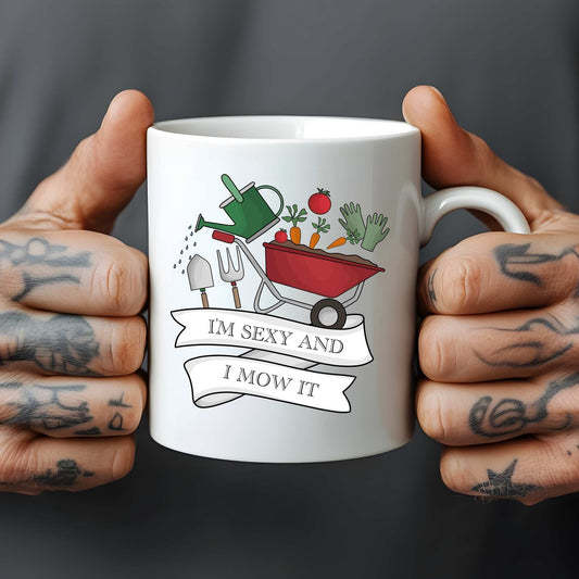 White mug held by tattooed hands with gardening-themed design and text.