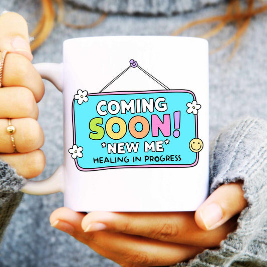 Person holding a mug with a colourful 'Coming Soon, healing in progress' design, featuring text and graphics.