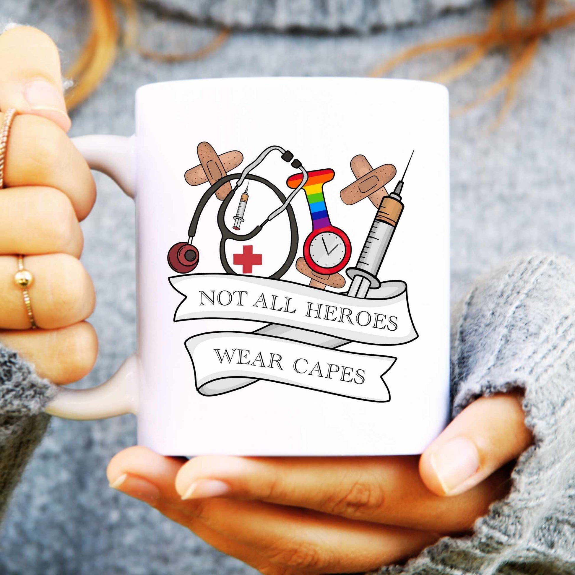 White mug held by hands with a design of medical symbols and text 'Not all heroes wear capes' on a grey background.