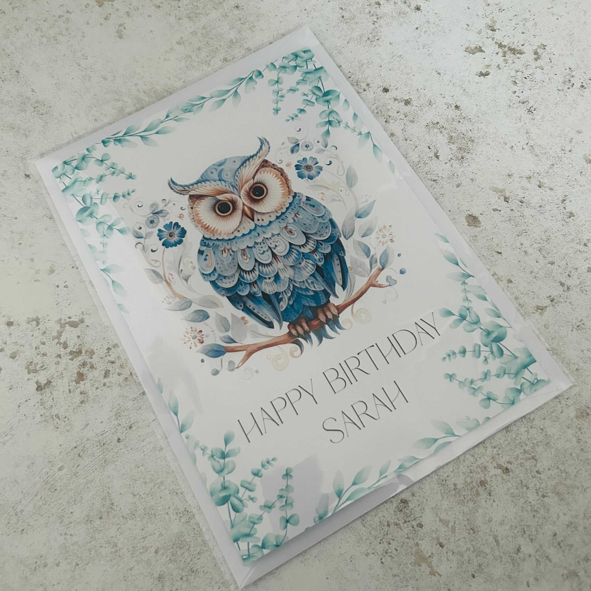Greeting card with a blue owl illustration and 'Happy Birthday Sarah' text on a textured surface.