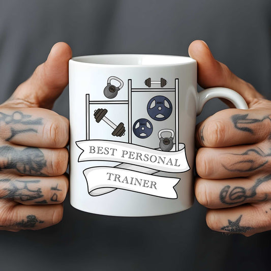Mug with gym-themed design and 'Best Personal Trainer' text held by tattooed hands.