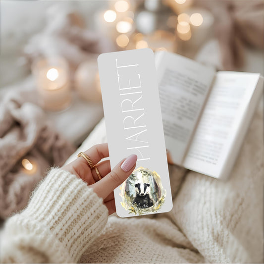 Hand holding a bookmark with 'HARRIET' and a badger design, blurred lights in the background