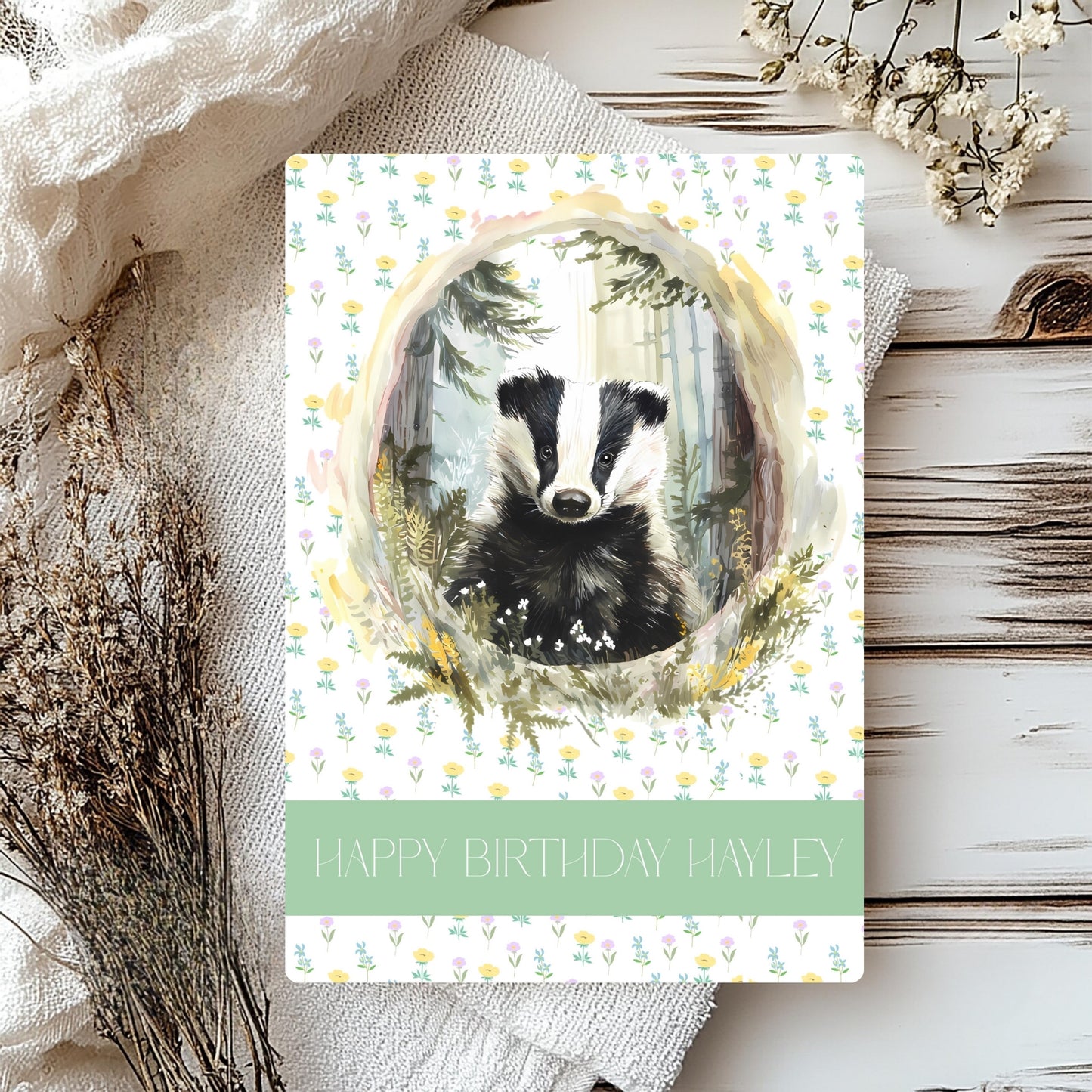 Greetings card with a badger illustration on a floral background, surrounded by dried flowers and fabric.