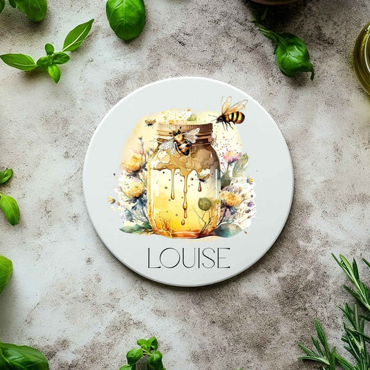 Round coaster with a design of a jar of honey, bees, and flowers on a concrete surface with basil leaves.