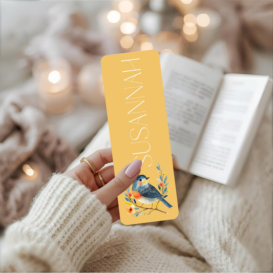 Hand holding a yellow bookmark with a bird illustration and 'SUSANNA' text, against a blurred cozy background.