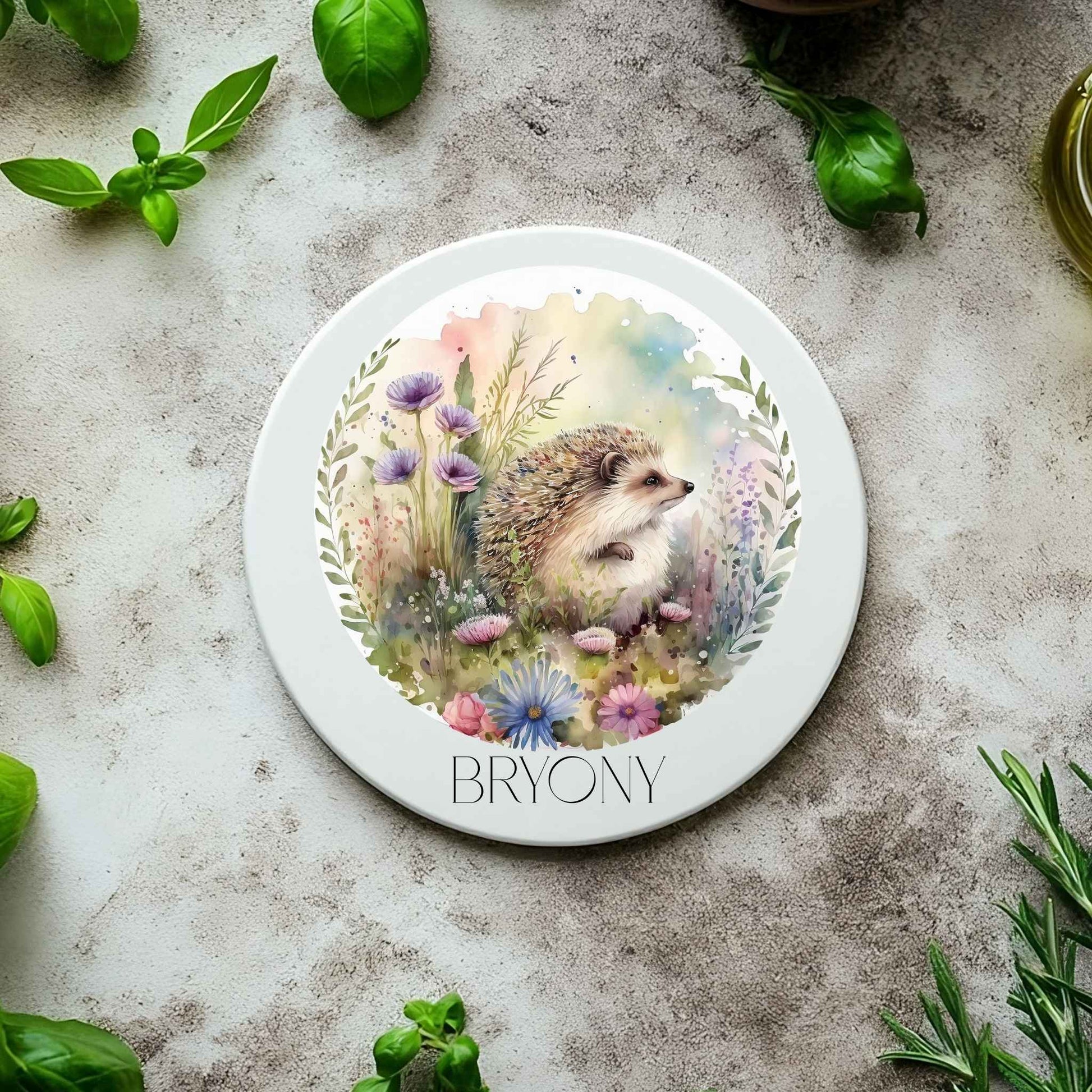 Ceramic coaster on a table surrounded by basil leaves with a hedgehog illustration and personalised.