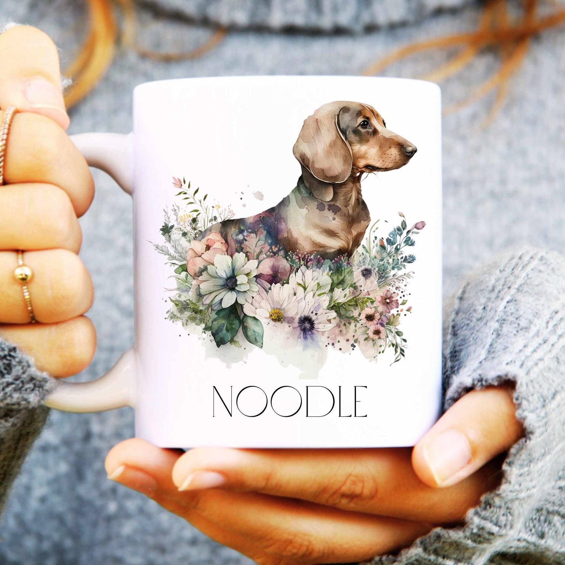 Person holding a mug with a dachshund and floral design, labelled 'Noodle'.