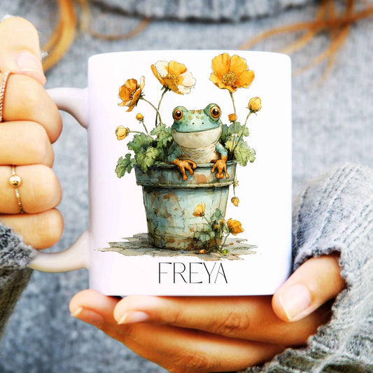 Person holding a ceramic mug with a design of a frog sat in a plant pot and personalised with a name.