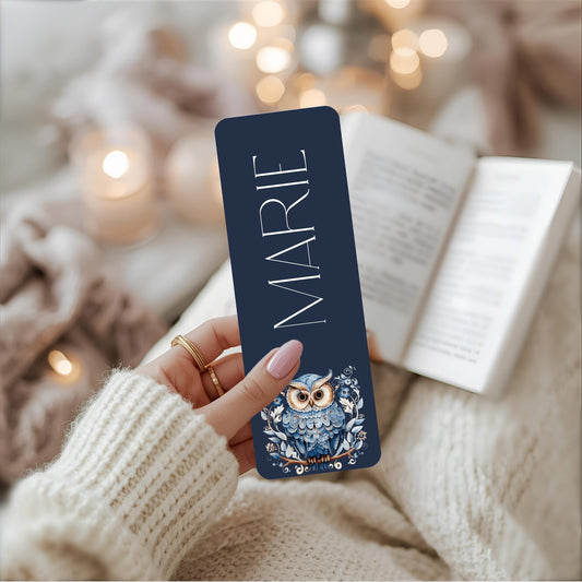 Person holding an owl bookmark with 'MARIE' on it, surrounded by soft lighting and cosy background