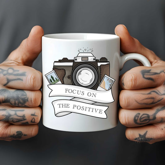 White mug with camera illustration and 'Focus on the Positive' text held by tattooed hands against a dark background