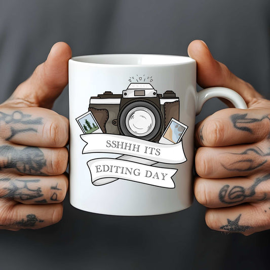 White mug held by tattooed hands with a camera graphic and text 'SSHHH ITS EDITING DAY' on a dark background