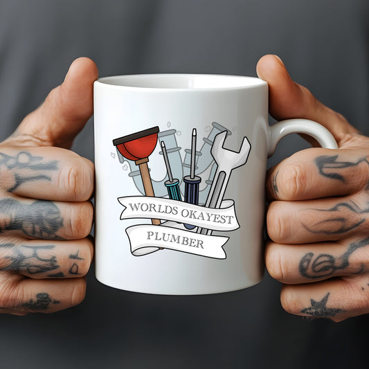 White mug held by hands with tattoos, featuring a graphic of plumbing tools and 'World's Okayest Plumber' text.