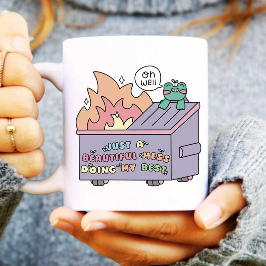 Mug with a cartoon design of a dumpster fire and text reads "just a beautiful mess doing my best", held by a person.