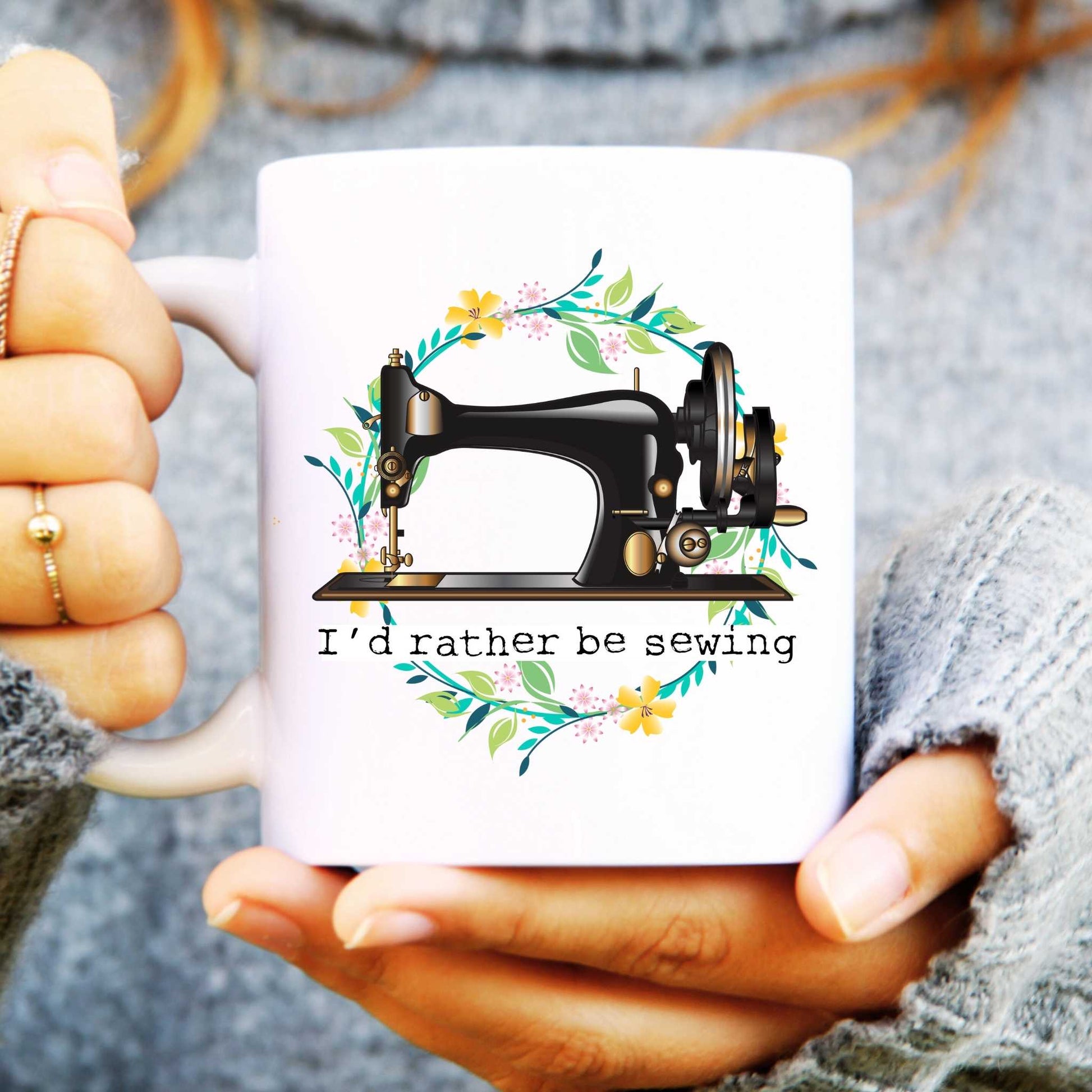 White mug with a sewing machine design and 'I'd rather be sewing' text, held by hands.