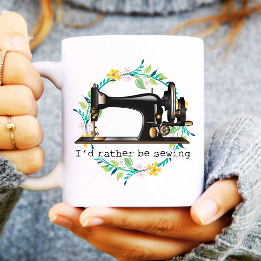 White mug with a sewing machine design and 'I'd rather be sewing' text, held by hands.