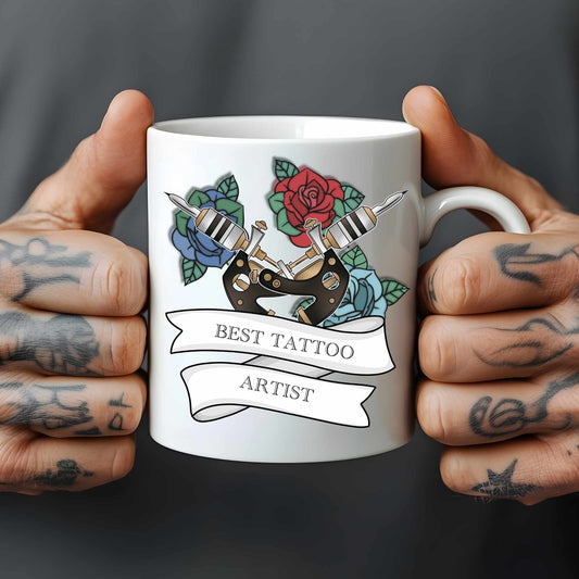 White mug held by tattooed hands with tattoo-themed design and 'Best Tattoo Artist' text.
