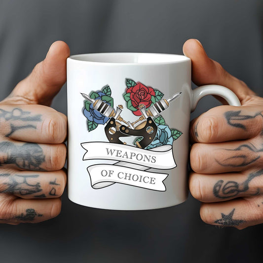 White mug held by tattooed hands with tattoo-themed design and 'Weapons of Choice' text.
