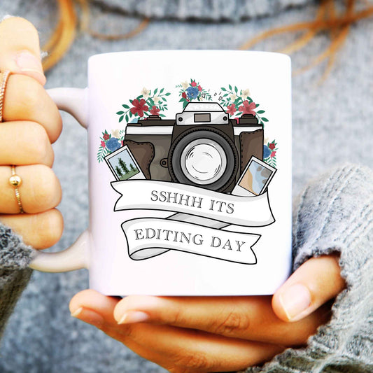 Person holding a mug with a camera design and text 'SSHHH ITS EDITING DAY'.